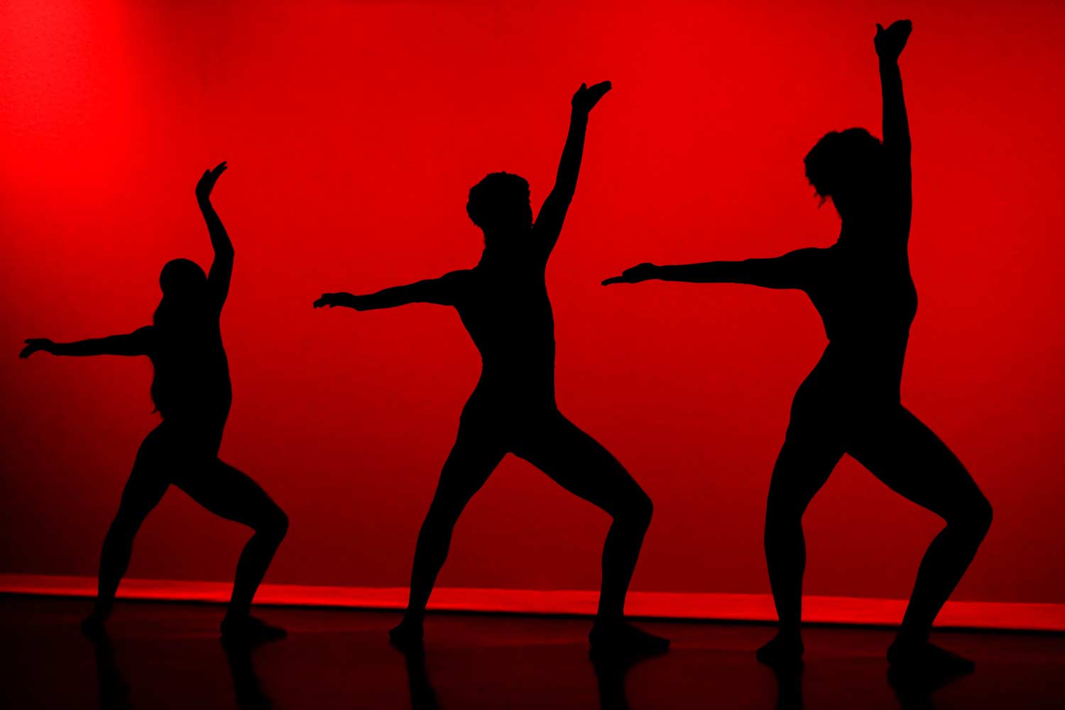 The silhouttes of three dancers on stage in front of a red background.
