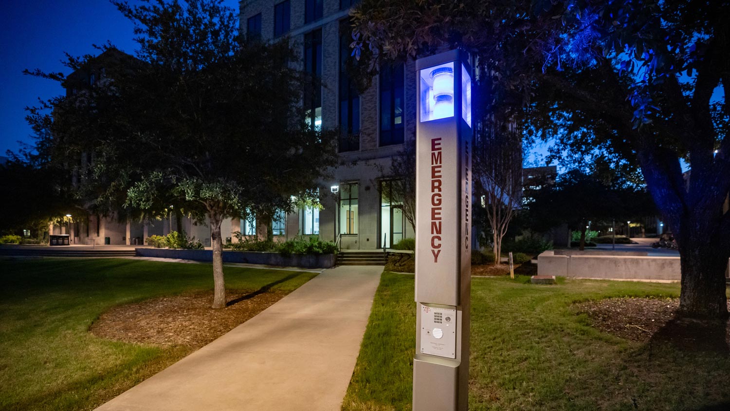 Silver emergency phone pole with blue light lit as dusk sets on campus