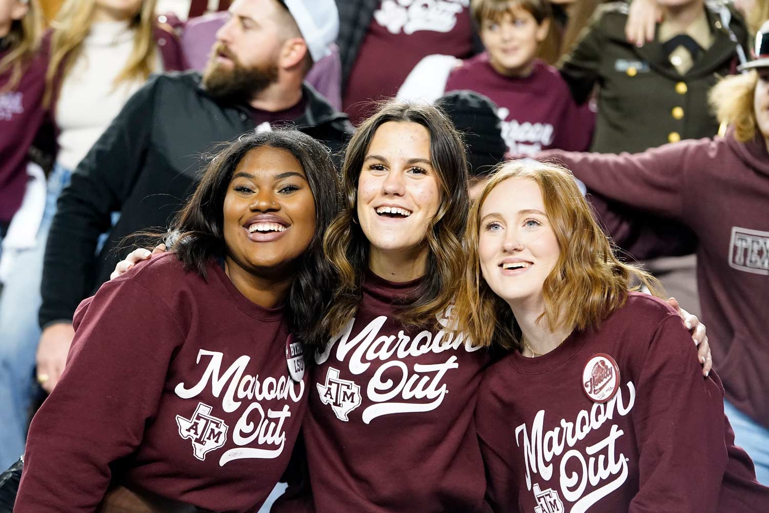 Three Aggies wearing maroon sweatshirts reading, "Maroon Out" pose at a football game.